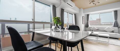 A modern living room with a large dining table, black chairs, a sofa, and a balcony with a view.