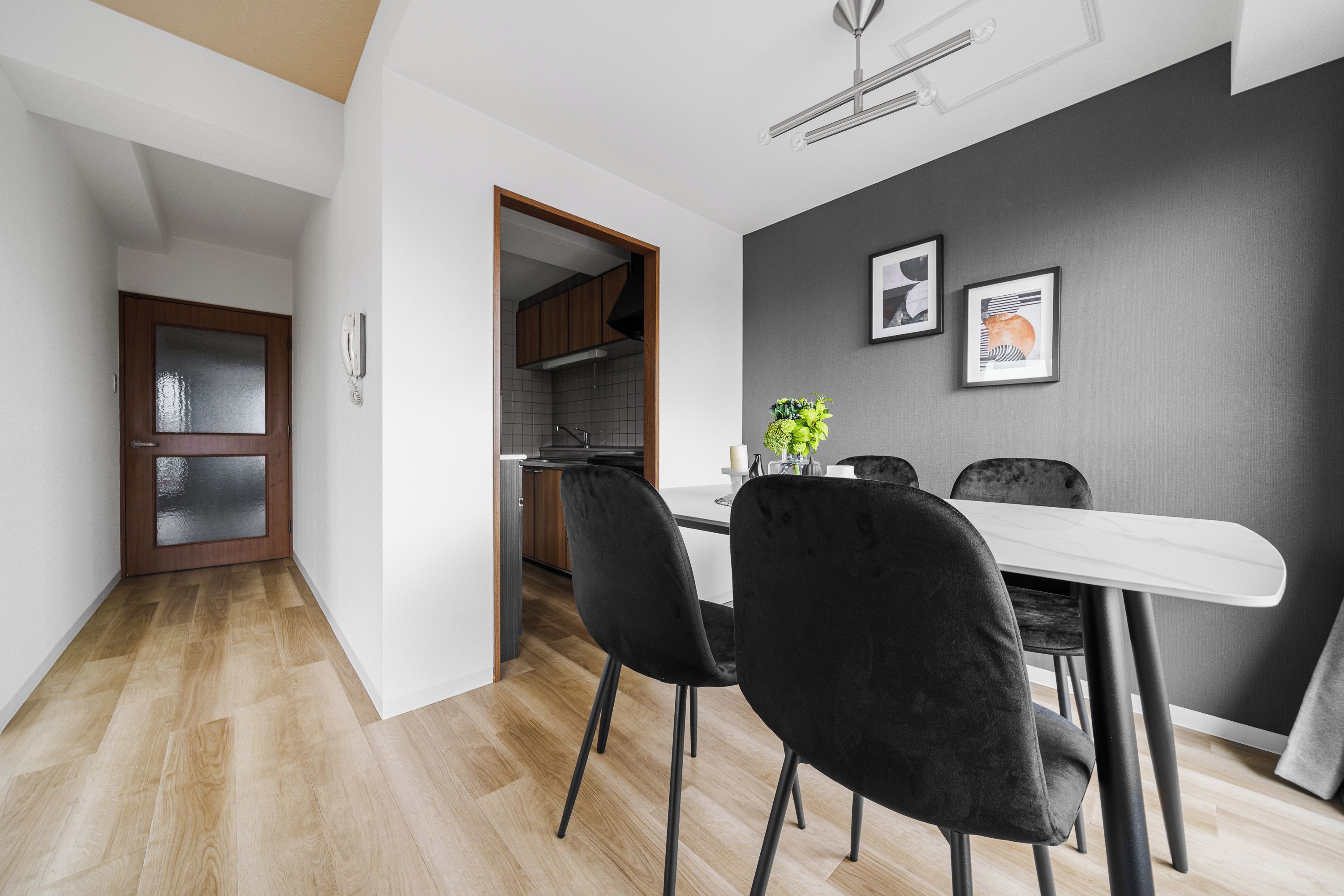 A modern dining area with a white table and black chairs, a kitchen in the background, and a grey wall with two framed pictures.