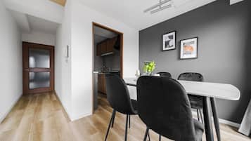 A modern dining area with a white table and black chairs, a kitchen in the background, and a grey wall with two framed pictures.