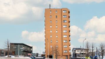 A tall brick building with multiple floors and windows, located in a snowy urban area with other buildings and a street in the foreground.