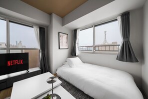 A hotel room with a bed, a TV displaying the Netflix logo, a framed picture on the wall, and a view of the cityscape through the windows.