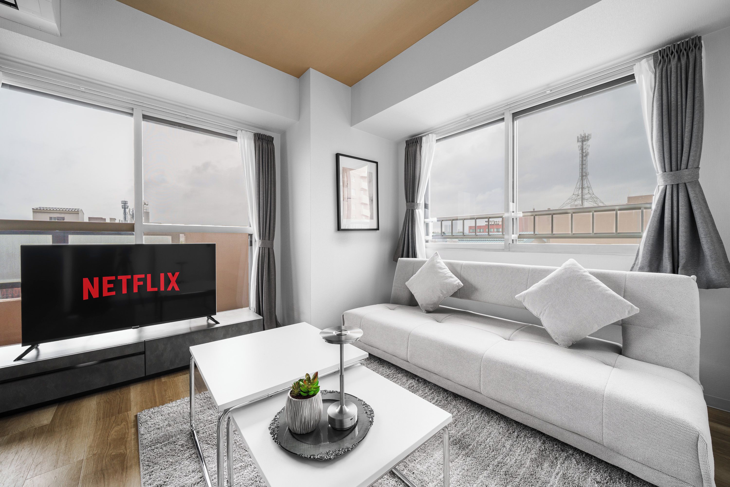 Ett modernt vardagsrum med en platt-TV, en vit soffa, ett soffbord med en växt och stora fönster med gardiner.