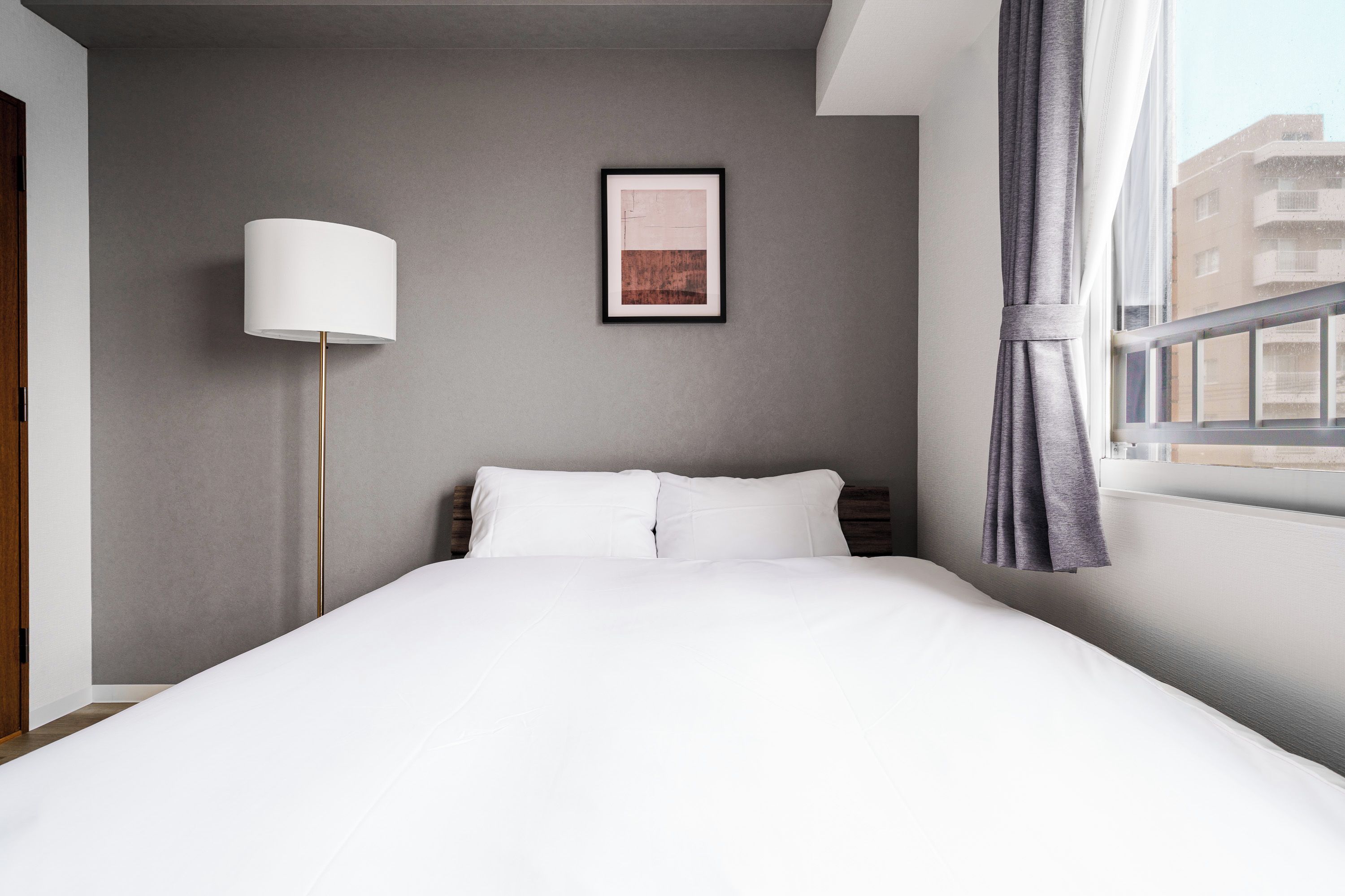 Ett hotellrum med en säng, en inramad tavla på väggen, en golvlampa och ett fönster med gardiner.