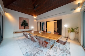 Uma zona de refeições moderna com uma mesa de madeira comprida, cadeiras de estrutura metálica e uma grande planta em vaso.