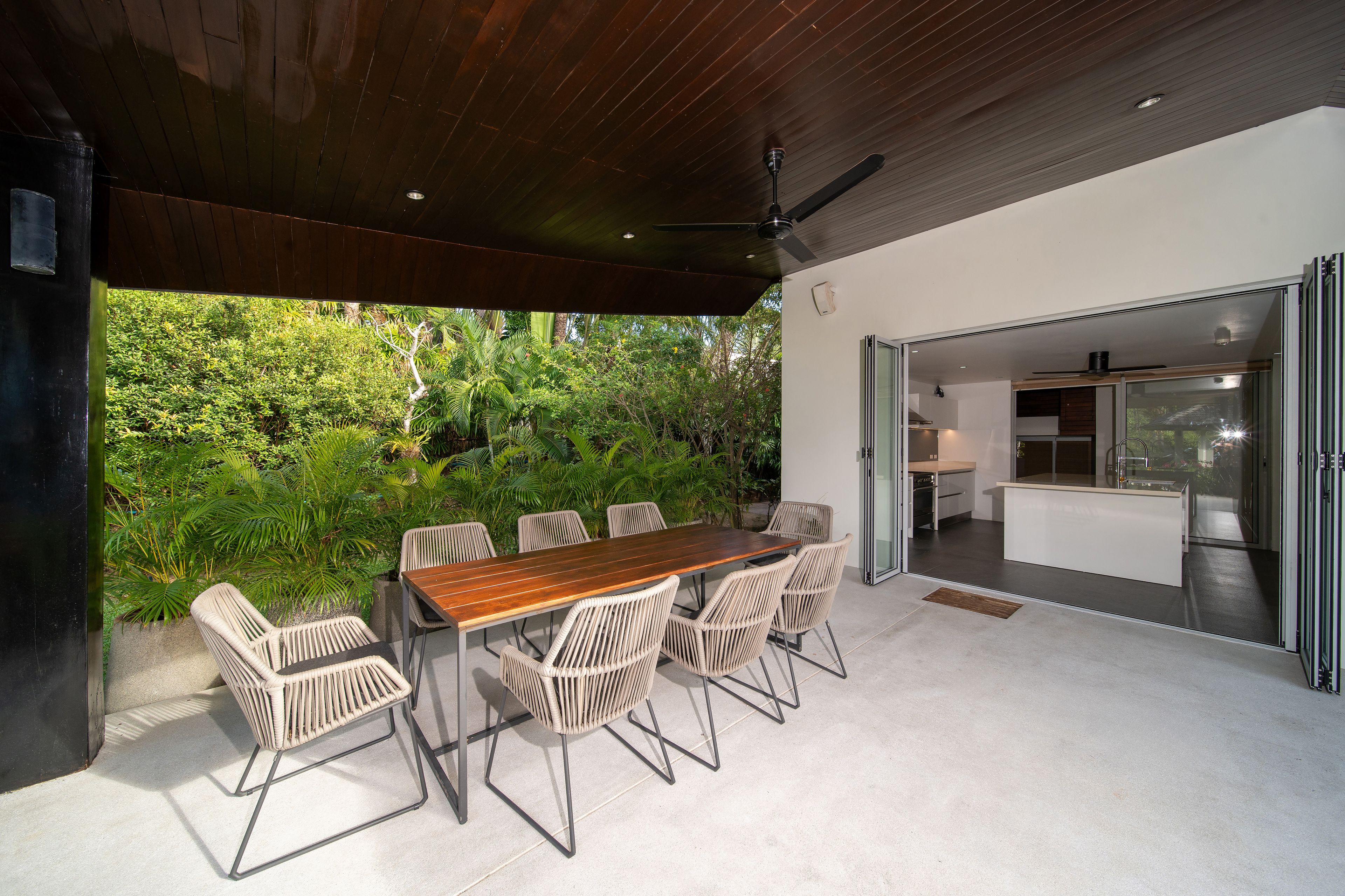 Un patio al aire libre con mesa de madera y sillas de mimbre, rodeado de abundante vegetación.
