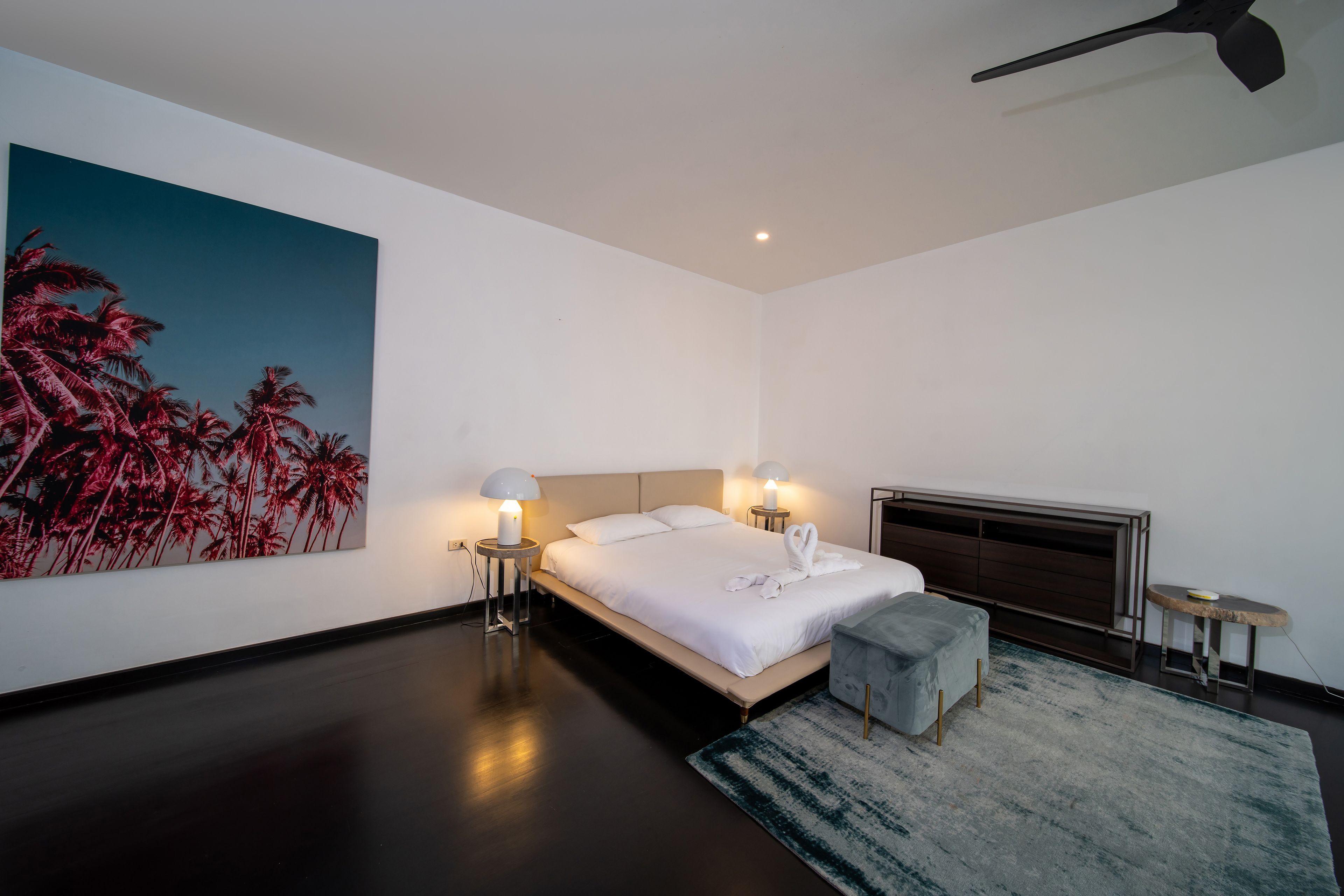 A modern bedroom with a large bed, bedside lamps, a painting of palm trees, a small table, and a bench.