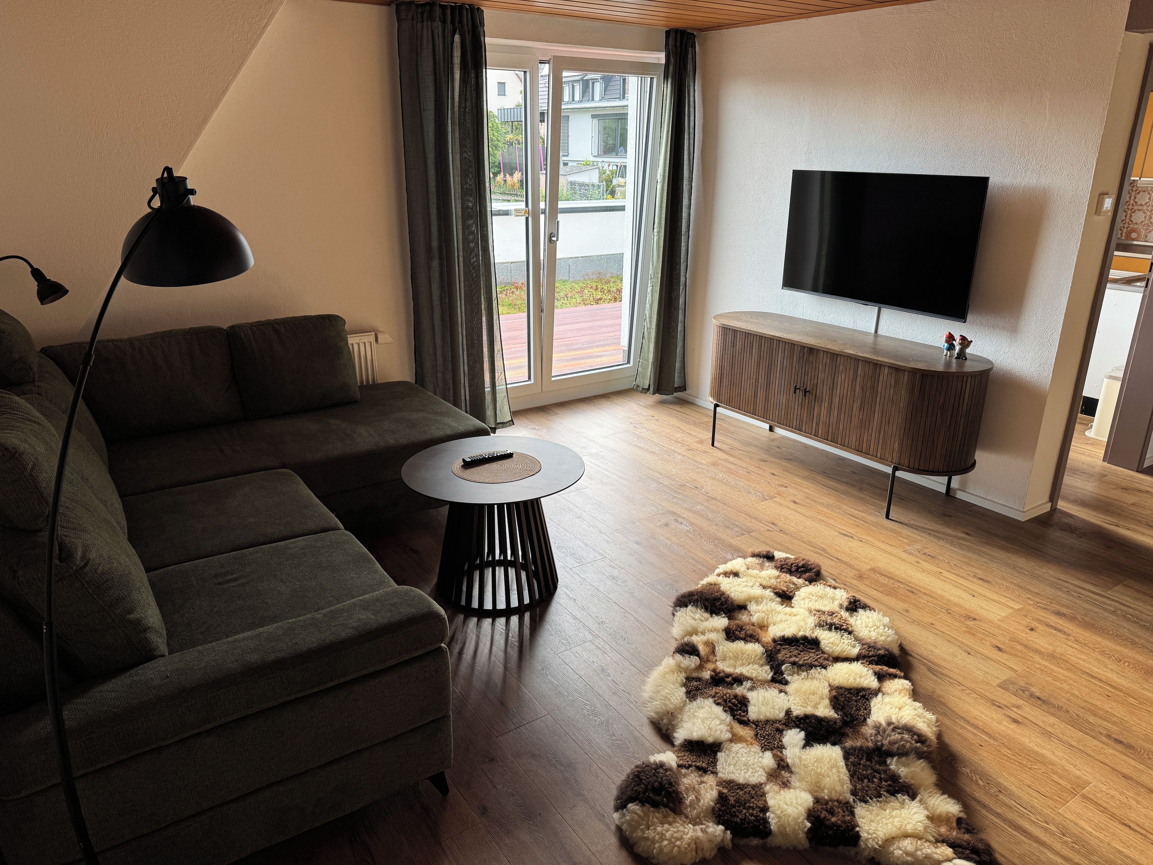 Ein modernes Wohnzimmer mit einem Ecksofa, einem kleinen runden Tisch, einem flauschigen Teppich, einem Fernseher auf einem Holzschrank und einem großen Fenster mit Vorhängen.