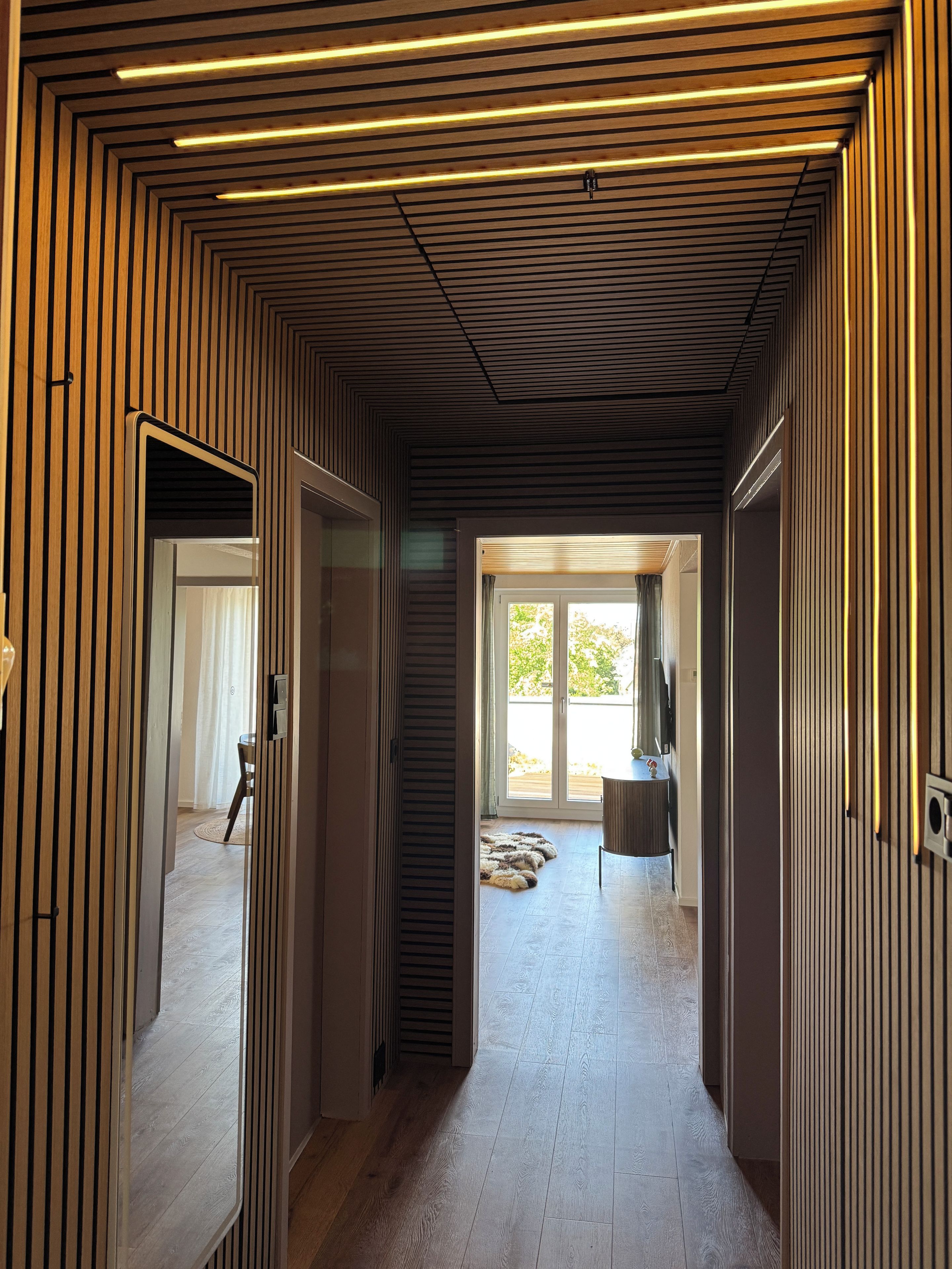 Ein modernes Interieur mit Wandgestaltung aus Holzlamellen, einer zentralen Treppe und Blick ins Freie.