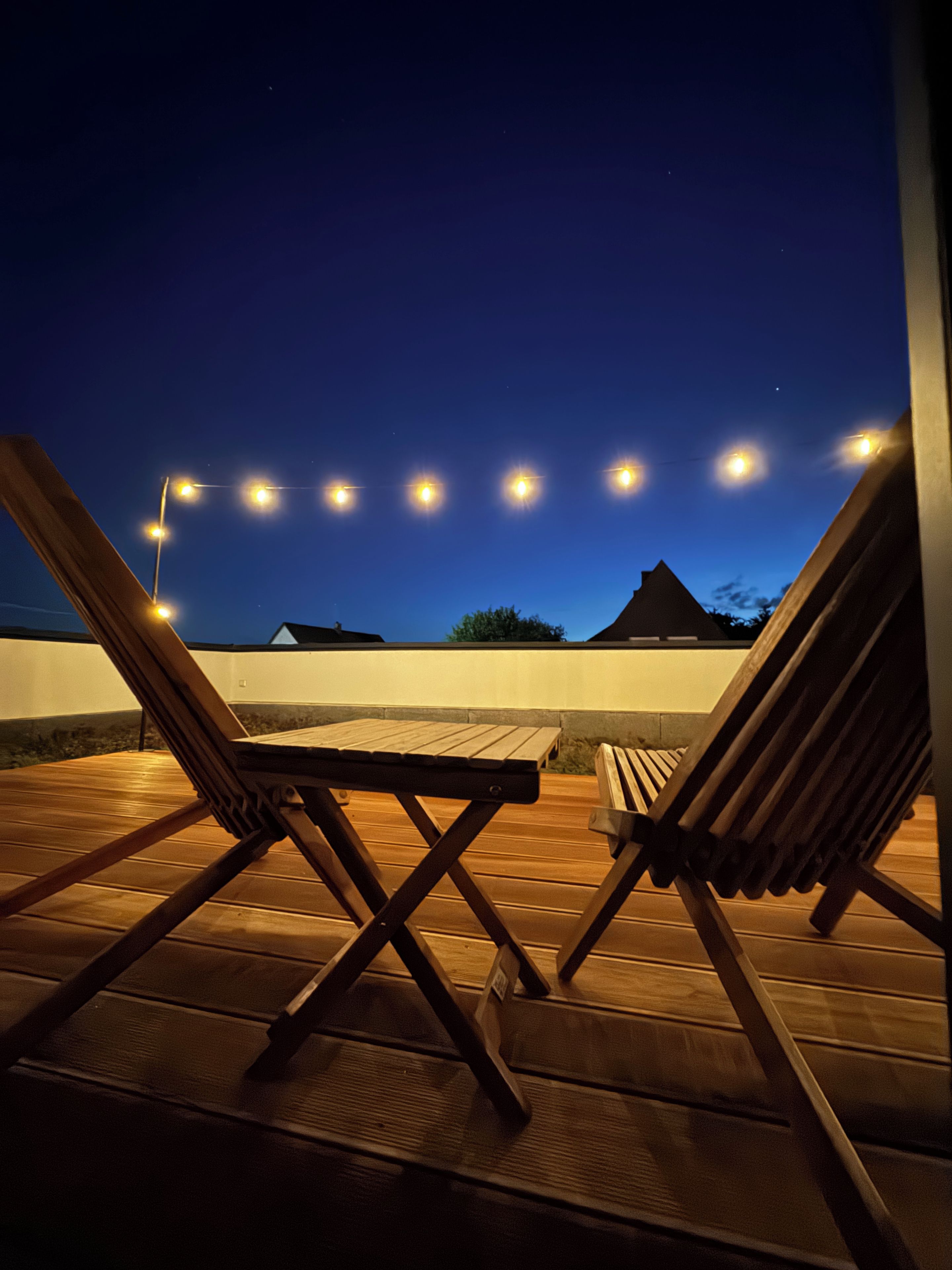 Eine Holzterrasse mit zwei Liegestühlen und einem Tisch, beleuchtet von Lichterketten vor einem Dämmerhimmel.