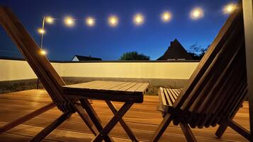 Eine Holzterrasse mit zwei Liegestühlen und einem Tisch, beleuchtet von Lichterketten vor einem Dämmerhimmel.