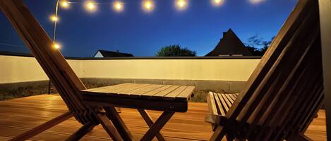 A wooden deck with two lounge chairs and a table, under string lights against a twilight sky.