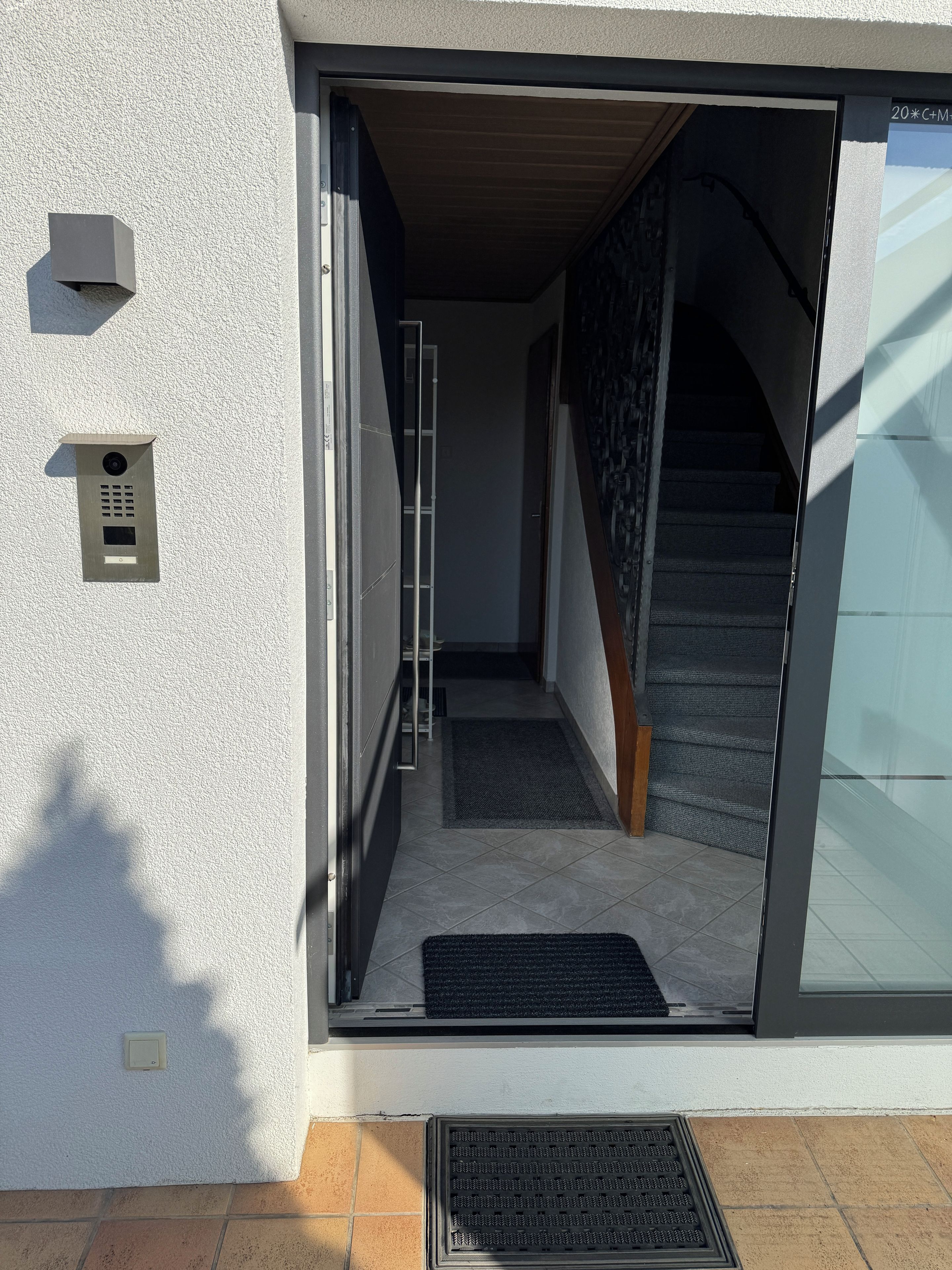 Ein moderner Hauseingang mit Glastür, einem Metall-Sprechanlage und einer sichtbaren Innentreppe.