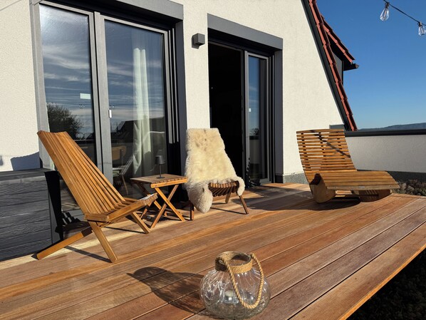 Una terraza de madera con sillones de descanso y una mesa, una puerta de vidrio que da al interior, y un cielo despejado.