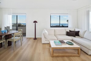 A modern living room with a white sofa, a wooden coffee table, and a dining area with a round table and chairs. - Design-Led Escape – Bondi & Tamarama – 2BR with Ocean Views (Bondi)