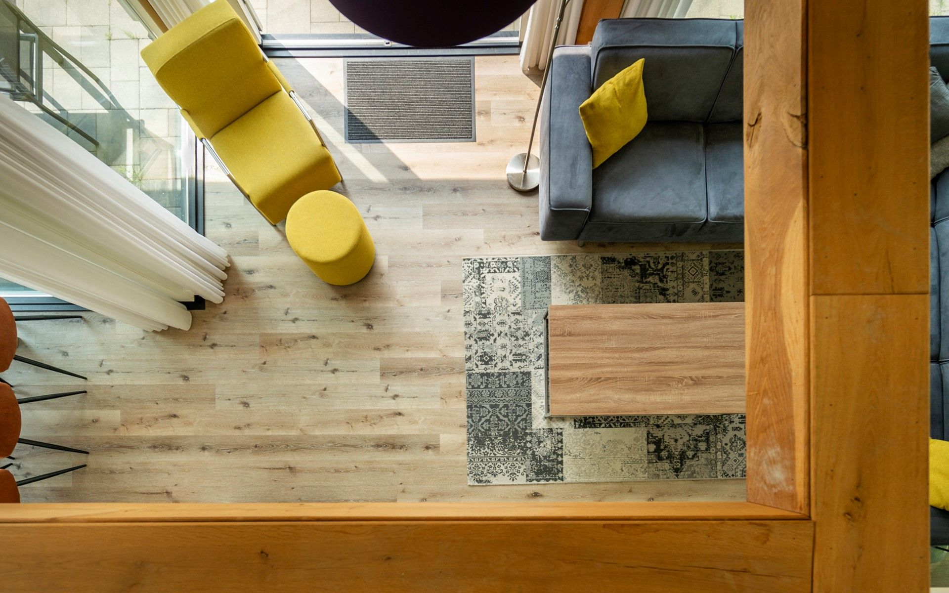 Een modern interieur met een houten vloer, een kamerkleed met patroon, een bank met kussens en een gele fauteuil.