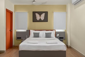 Un dormitorio con una cama, dos mesitas de noche, una lámpara de pared, una obra de arte con mariposas enmarco y una ventana con persianas.