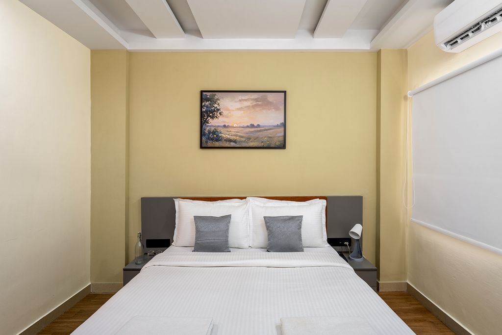 Habitación de hotel con cama, dos mesitas de noche, un velador, aire acondicionado de pared y un cuadro en la pared.