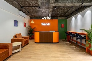 Zona de recepción con un mostrador de madera, paredes naranja y una pared de acento verde. Hay áreas de estar con sillas y mesas, y plantas en macetas.