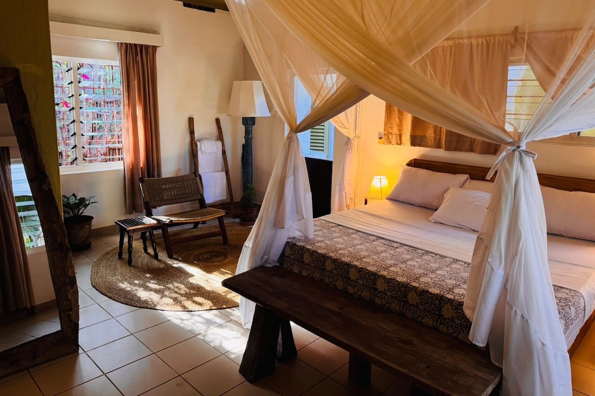 A bedroom with a bed, a bench, a chair, a lamp, and a window with curtains.