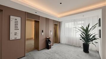 A hotel lobby with a reception desk, a directory on the wall, a potted plant, and a large window with sheer curtains.