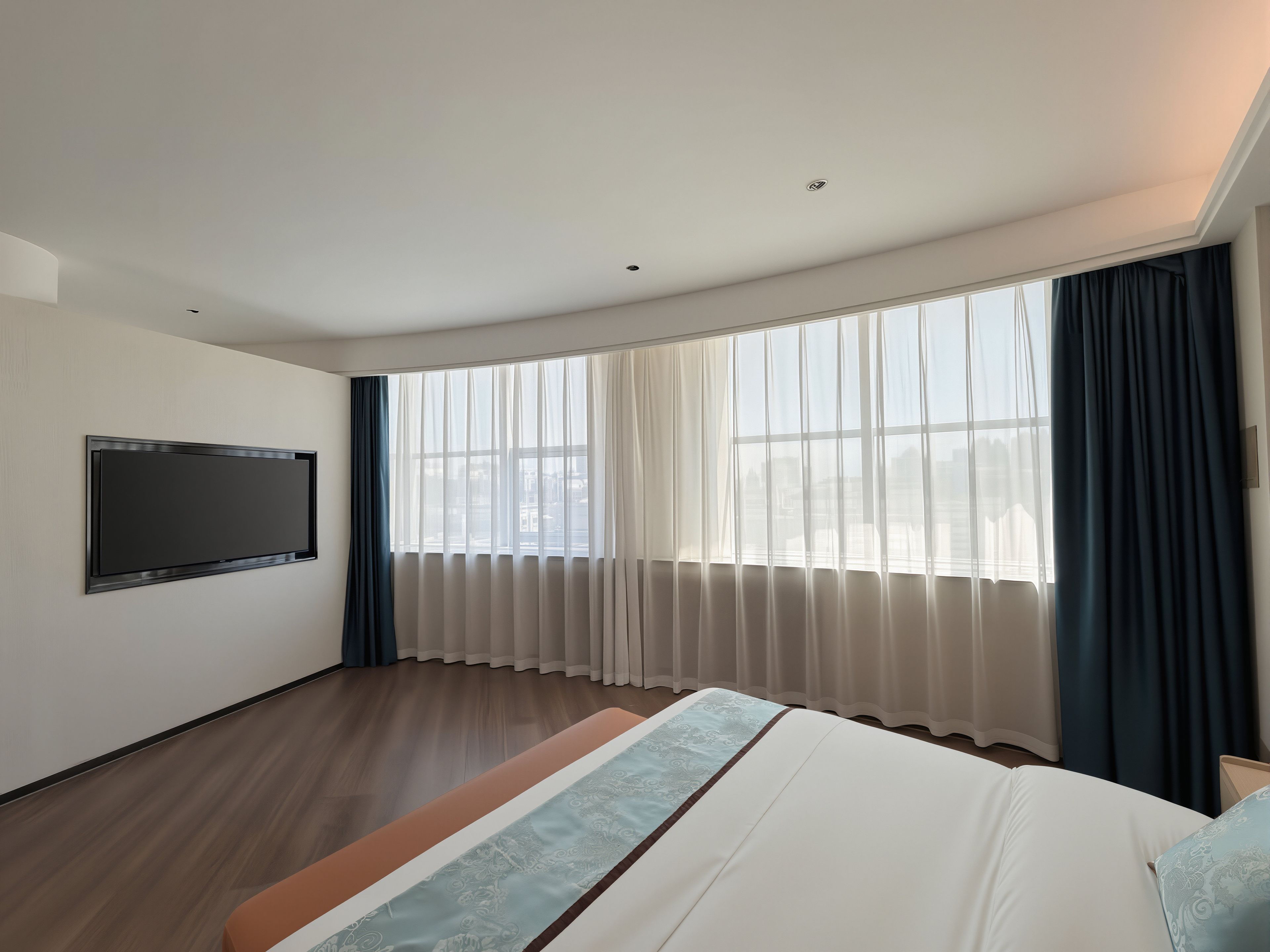 A hotel room with a large bed, a flat-screen TV mounted on the wall, and a large window with sheer curtains.