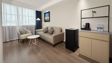 A modern living room with a sofa, armchair, and coffee table. There is a small kitchen area with a refrigerator and a shelf with water bottles.