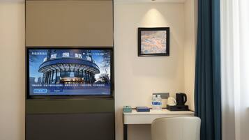 A hotel room with a bed, a desk with a chair, a television mounted on the wall, a framed picture, and a window with curtains.