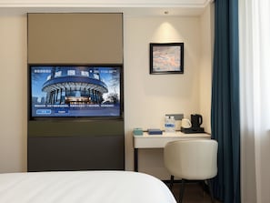 Habitación de hotel con una cama, un escritorio con silla, un televisor en la pared, un cuadro enmarcado y una ventana con cortinas.