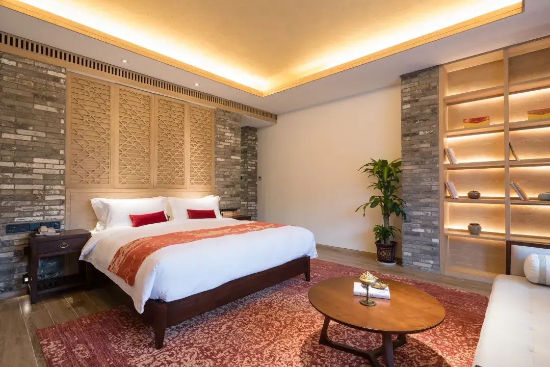 A modern hotel room with a large bed, a bedside table, a round coffee table, and a bookshelf.