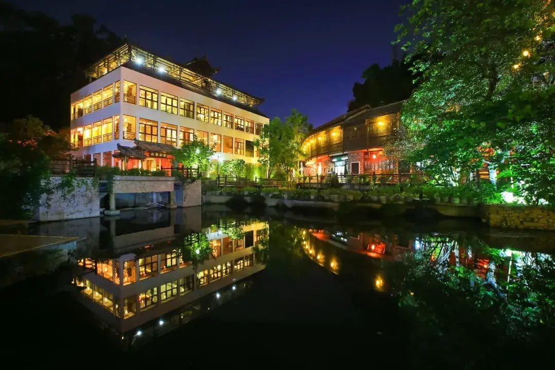 Un edificio de varios pisos con su reflejo en el agua, rodeado de vegetación e iluminado con luces artificiales.