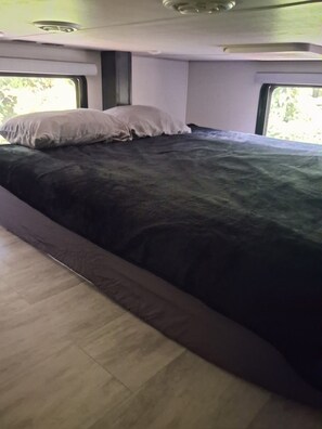 A bed with a dark cover and pillows, a window with a view of greenery, and a ceiling with a light fixture.