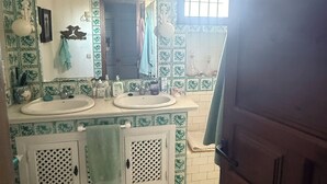 Een badkamer met twee wasbakken, een toilet en een spiegel. Aan de rechterkant is een raam met tralies.