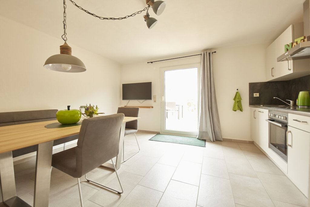 A modern kitchen with a dining area, a TV mounted on the wall, and a view through a glass door.