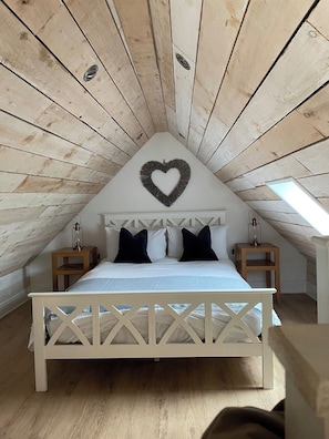 Un dormitorio con techo inclinado, una cama con estructura blanca, dos mesitas de noche y una decoración con forma de corazón en la pared.