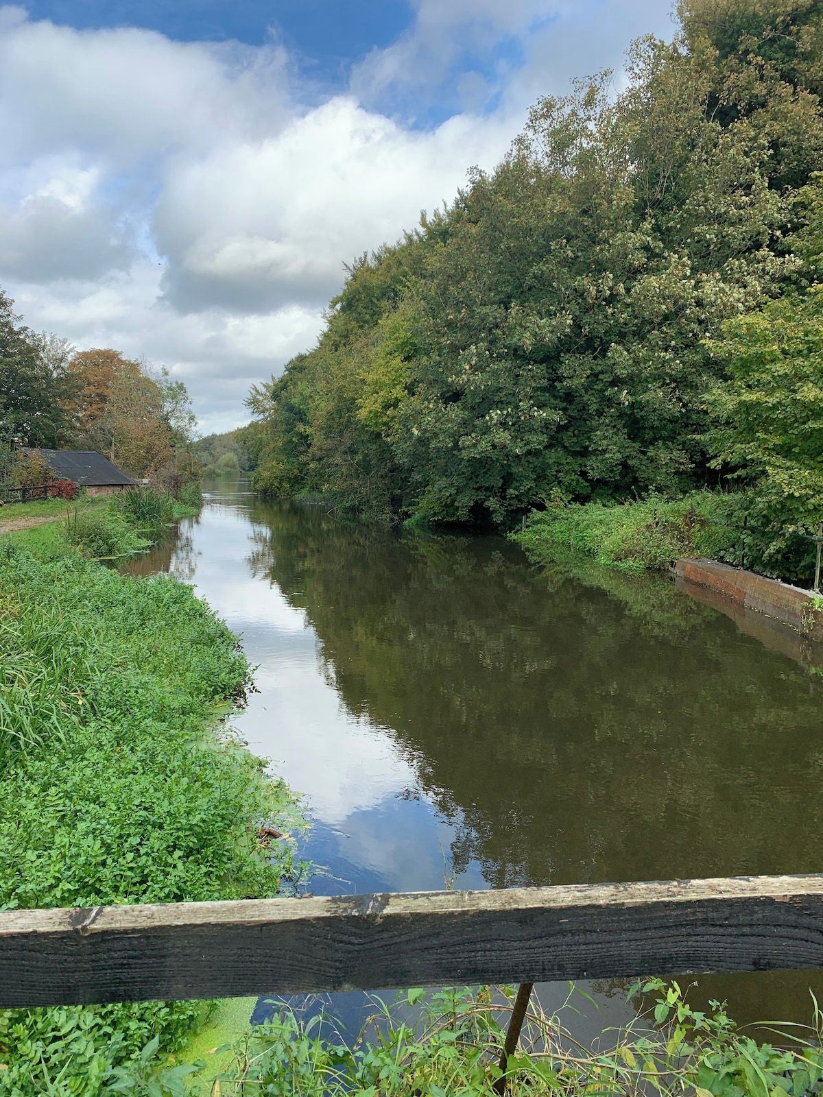 A calm river with dense greenery on both sides, a wooden fence in the foreground, and a partly cloudy sky.