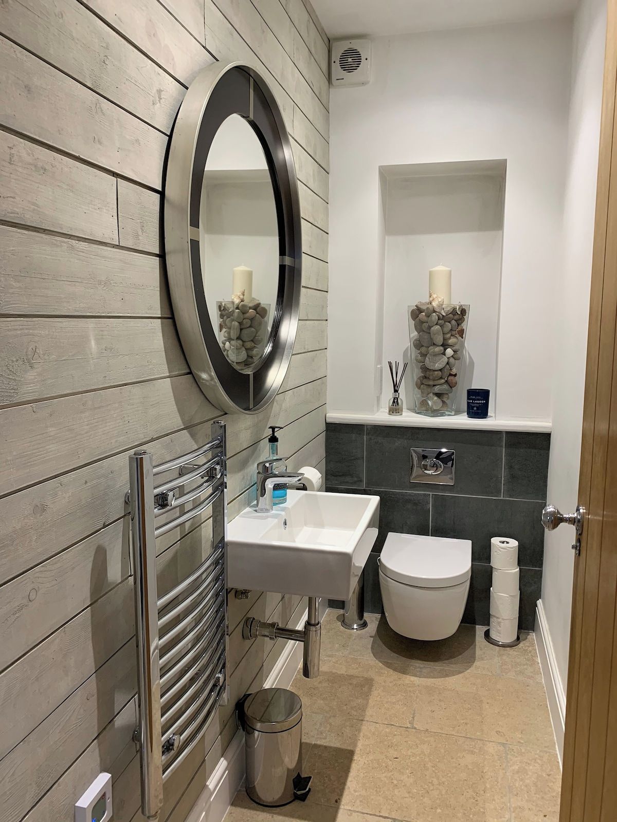 Een moderne badkamer met een houten wand, een ronde spiegel, een witte wasbak, een toilet en een kleine prullenbak.