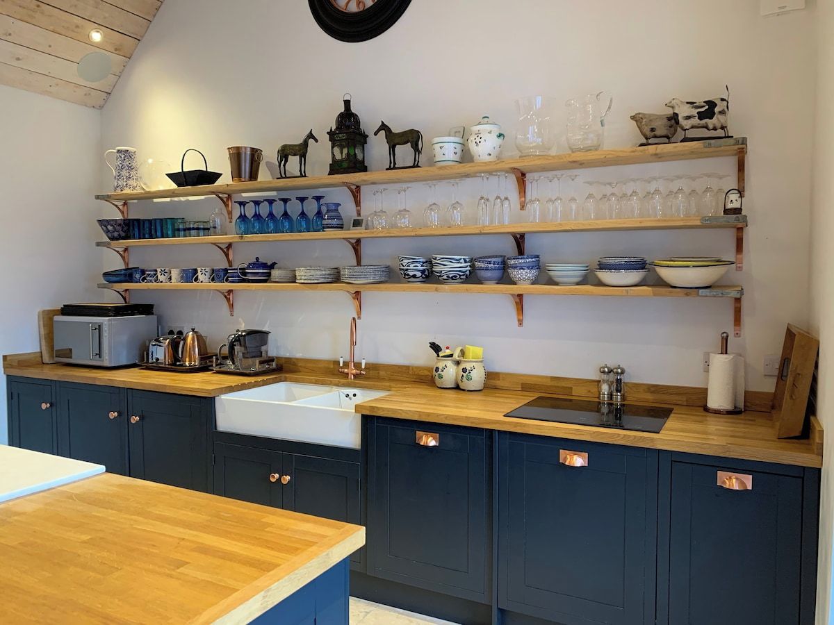 A kitchen with dark cabinets, copper handles, a wooden countertop, and shelves with various glassware and decorative items.