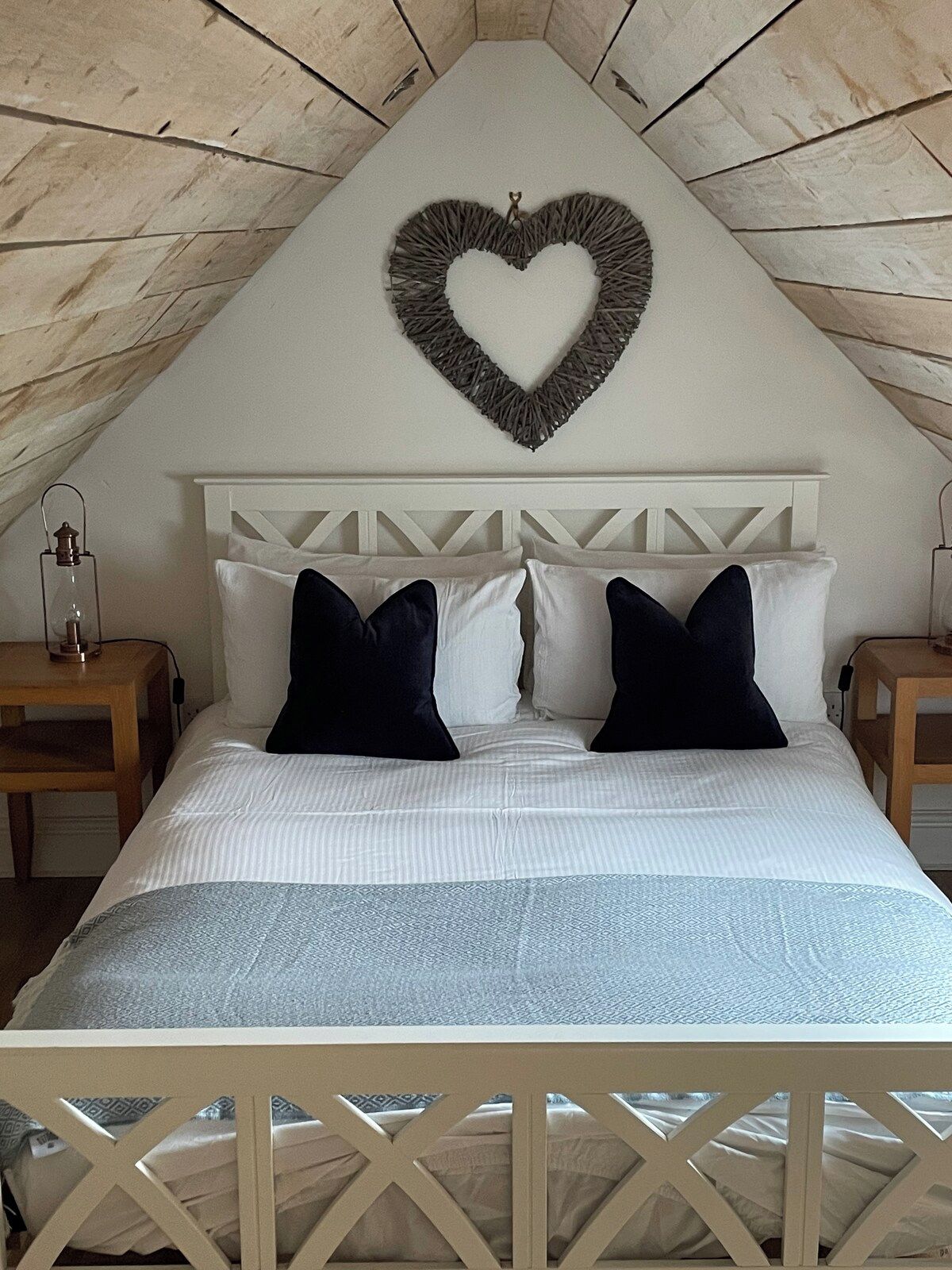 Een slaapkamer met een schuine plafond, een hartvormige decoratie aan de muur, een bed met witte en donkere kussens en een houten nachtkastje met een lamp.