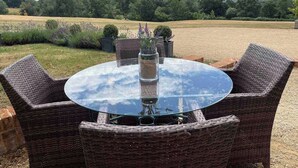 Et rundt glastræbord med fletstole, omgivet af et grusbed med planter og udsigt til et mark og træer.
