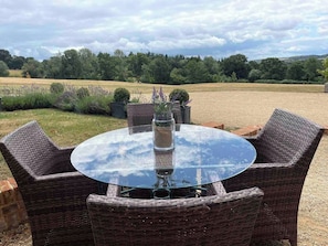 Mesa de vidro redonda com cadeiras de vime, cercada por uma área de cascalho com plantas e vista para um campo e árvores.