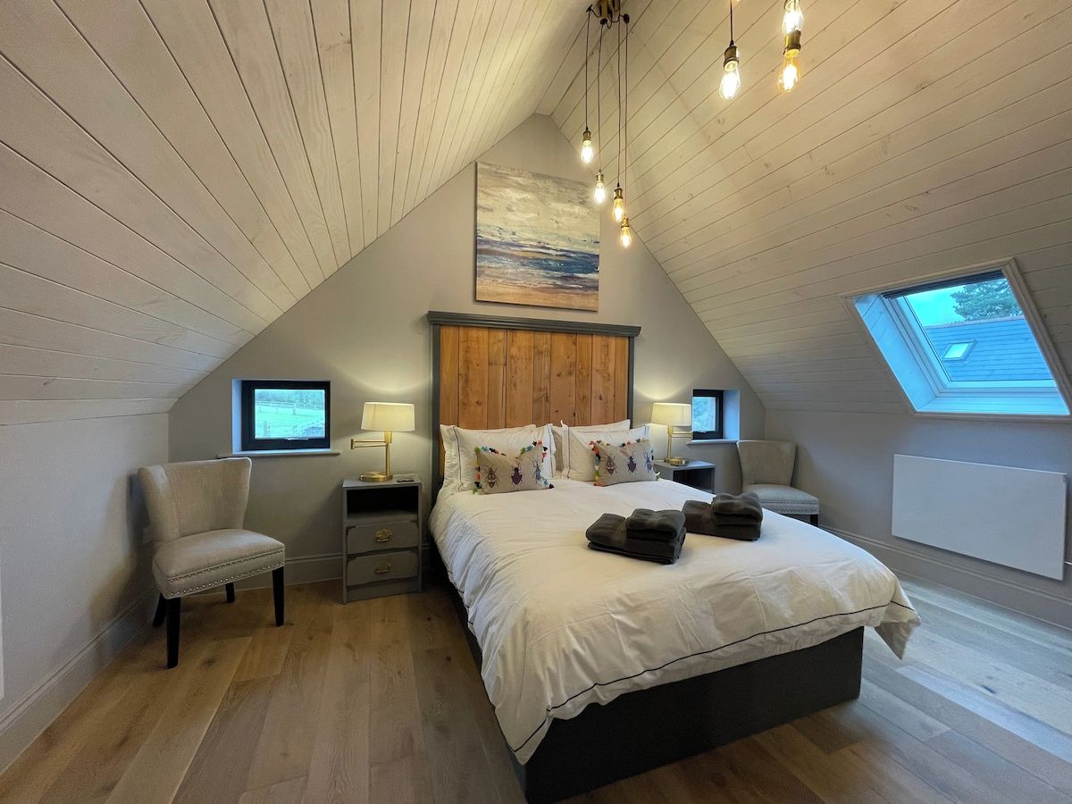 Un dormitorio con techo inclinado, una cama con almohadas, una silla, mesitas de noche y un cuadro en la pared.