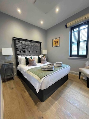 Quarto com uma cama grande, cabeceira com padrão geométrico, mesa de cabeceira com abajur e janela com persianas.