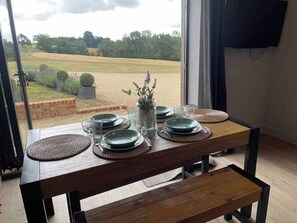 Mesa de jantar posta com pratos, copos e um vaso de flores, posicionada perto de uma janela com vista para um campo.