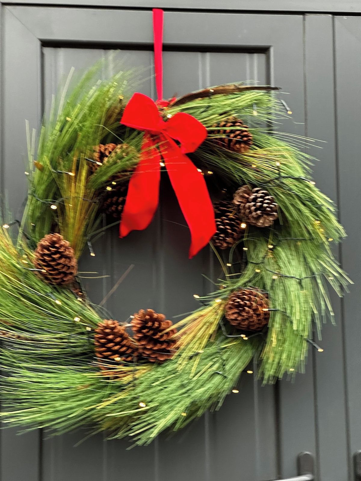 Une couronne décorée de branches de pin, de pommes de pin et d’un ruban rouge, posée sur une porte grise.