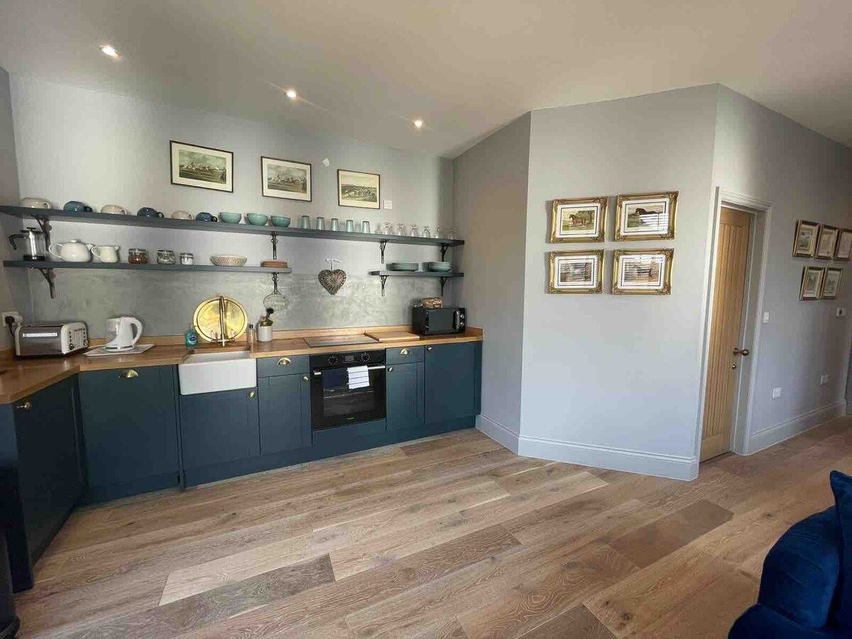 A modern kitchen with dark cabinetry, a wooden countertop, and a built-in oven. There are shelves above the counter holding various kitchenware and framed pictures on the wall.