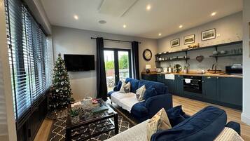 A living room with a blue sofa set, a coffee table, a Christmas tree, and a kitchen area with a window and shelves.