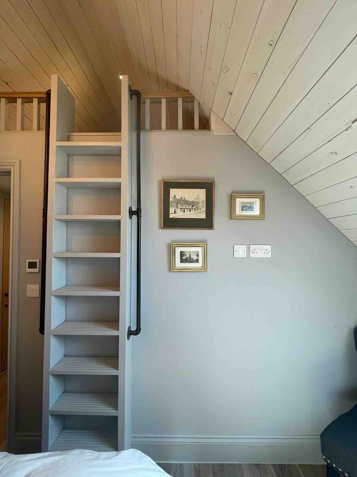 Una habitación con una escalera de madera que sube a una zona de altillo, una cama y dos cuadros en la pared.