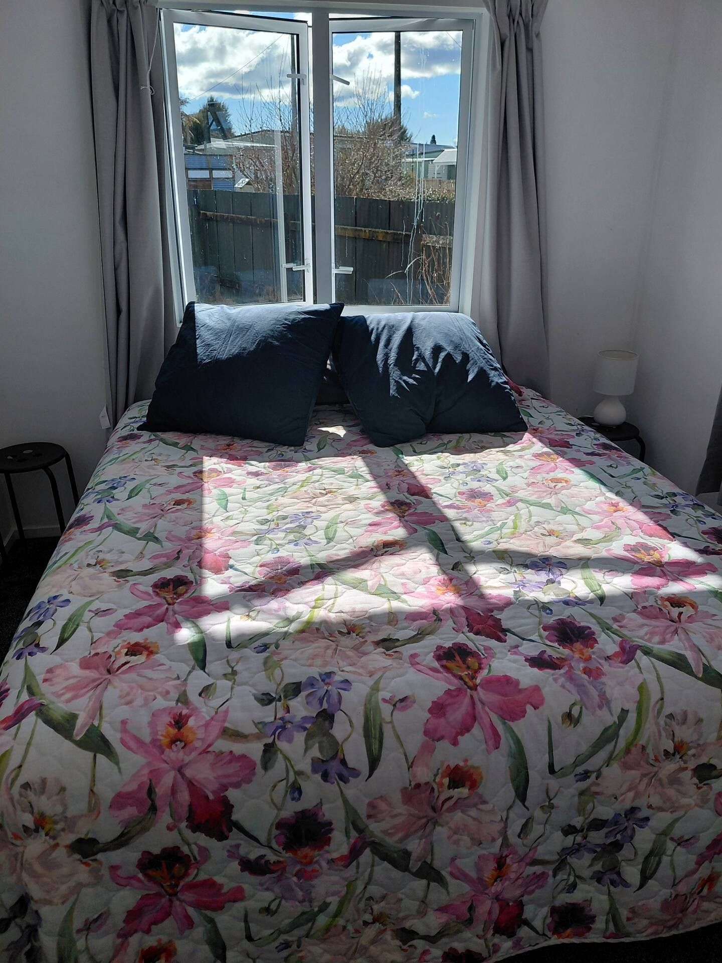 A bed with a floral bedspread, two dark pillows, a window with curtains, and a lamp on a bedside table.