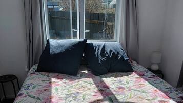 A bed with a floral bedspread, two dark pillows, a window with curtains, and a lamp on a bedside table.