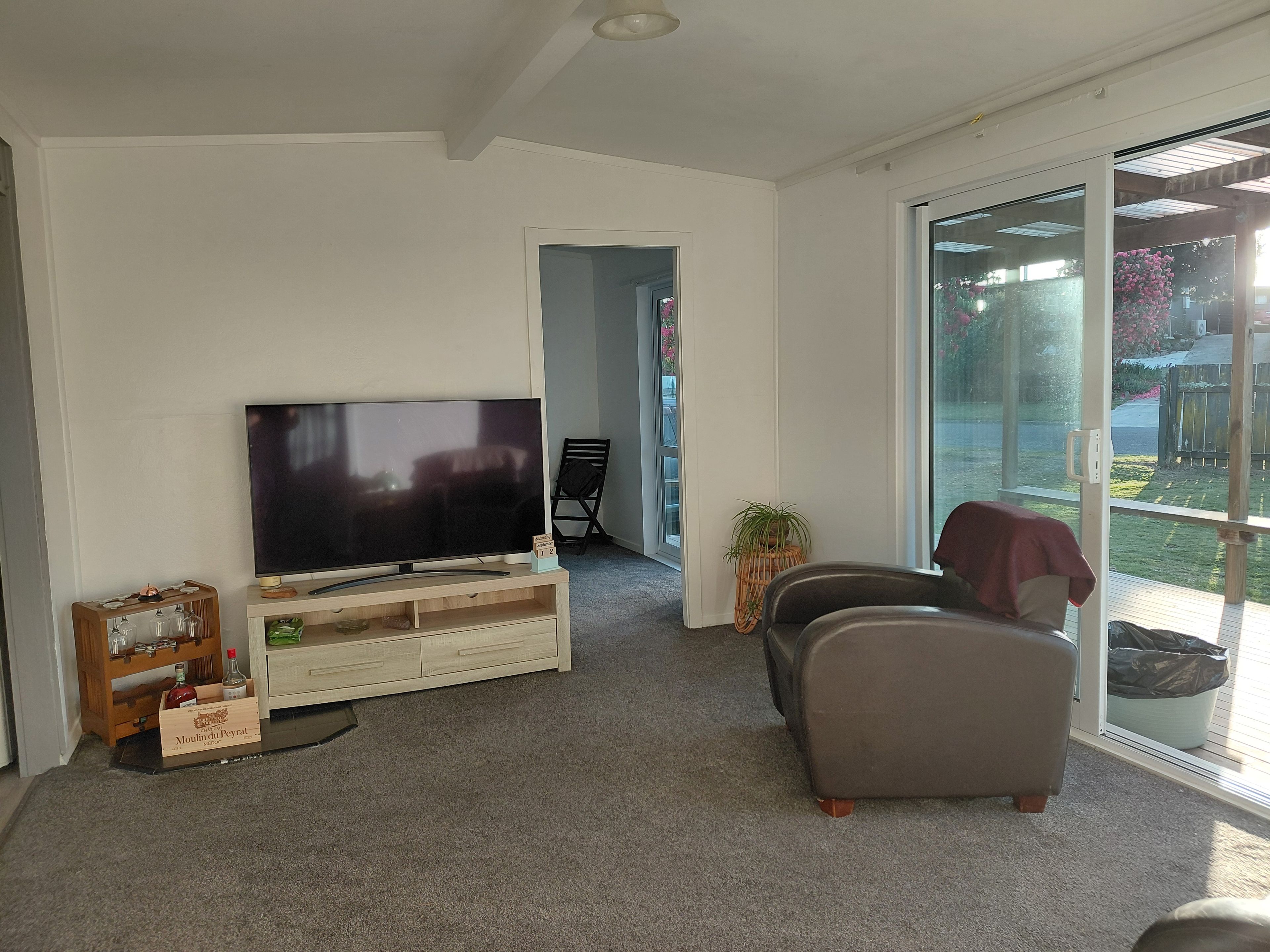 En stue med et fladskærms-tv på et træstel, en sofa, et lille bord med glas og flasker, samt en skydedør til en have.